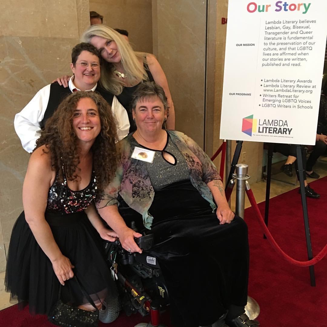Photograph of 4 women dressed up in fancy clothes. Next to them is a sign for the Lambda Literary Awards.