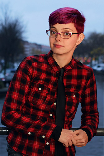 Photograph of Corinne Duyvis, a lightskinned, 20ish person with short red hair, metal framed glasses wearing a red and black plaid shirt and a black tie. A blurry outdoor background of water, boats and trees is behind.