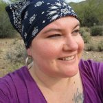 Photograph of Naomi Ortiz, a Mestiza (Indigenous/Latina/White) disabled woman. She wears a dark blue and white bandana on her head, large silver earrings, and a purple blouse. A tattoo on her left chest shows the word "Voice" in cursive writing.